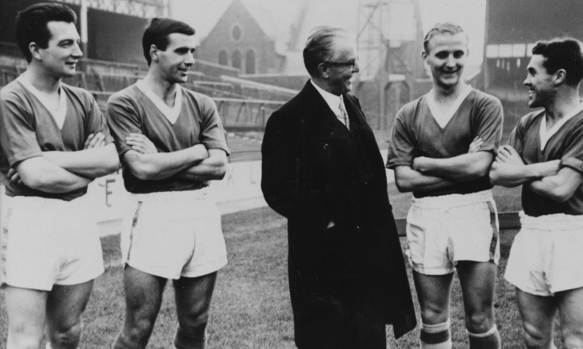 A black and white photograph of four male footballers stood around Sir John Moores on a football pitch