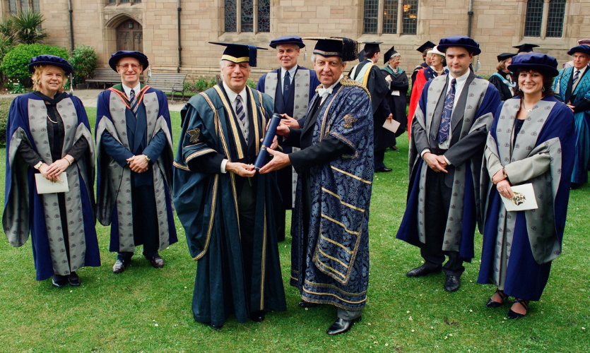 A group of LJMU staff wearing graduation caps and gowns with John Moores Junior stood at the front being passed a scroll