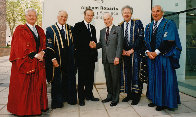Aldham is dressed in a grey suit and is shaking hands with another man, surrounded by other men dressed in long robes stood outside of the Adham Robarts library building .
