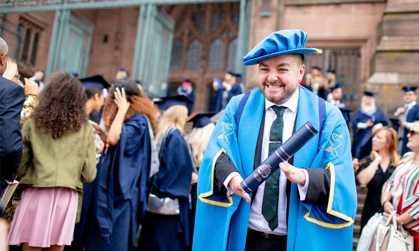 Alex is dressed in a blue cap and gown stood outside of the Anglican Cathedral, his hand and arm disabilities can be seen as he holds a scroll in his hands.
