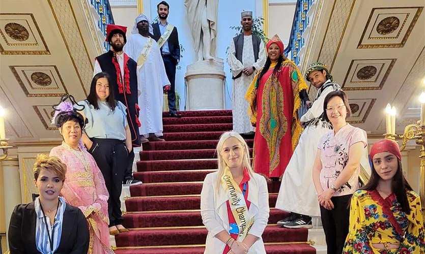 An image of Alexandra wearing a gold sash across her body that says the words Community Champion, she is stood on a set of red steps surrounded by other people wearing traditional cultural dress who also have gold sashes over their outfits