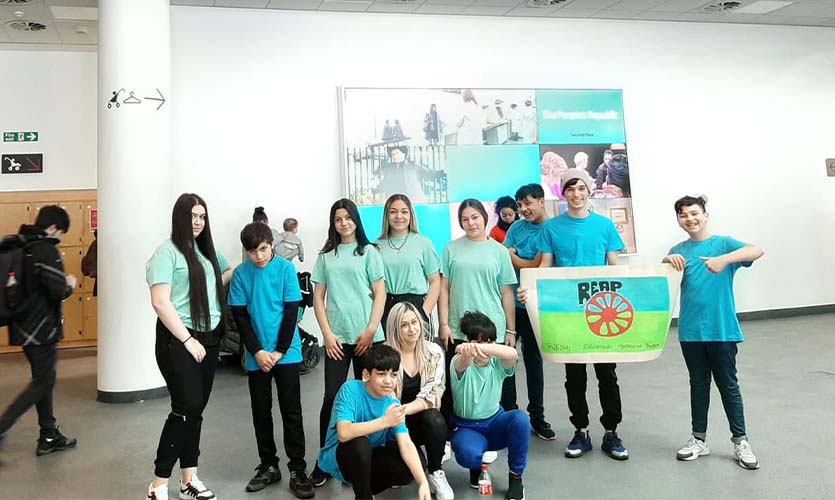 An image of Alexandra crouched on the floor surrounded by a group of young people all wearing green t-shirts, they are stood in a large white painted atrium of a Liverpool museum building