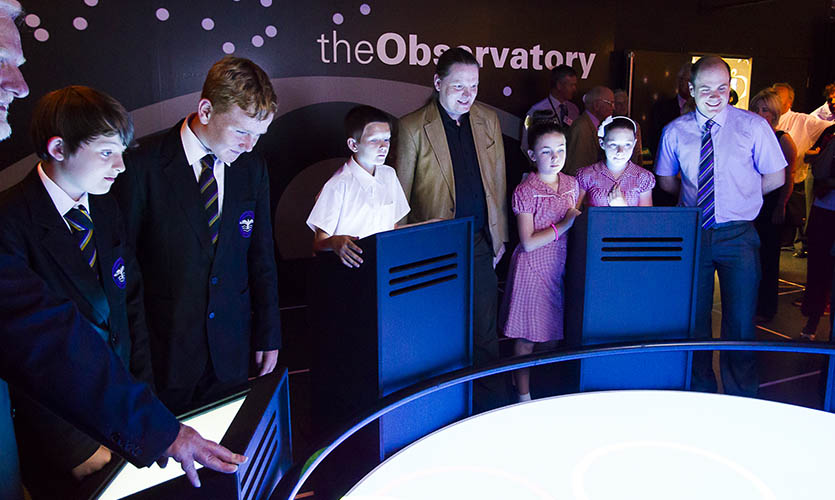 Andy stood between a group of schoolchildren who are looking at simulations of the universe
