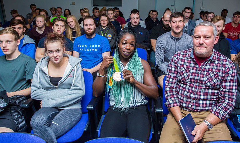 Anyika sat down holding her Olympic medal in her hands surrounded by LJMU staff and students