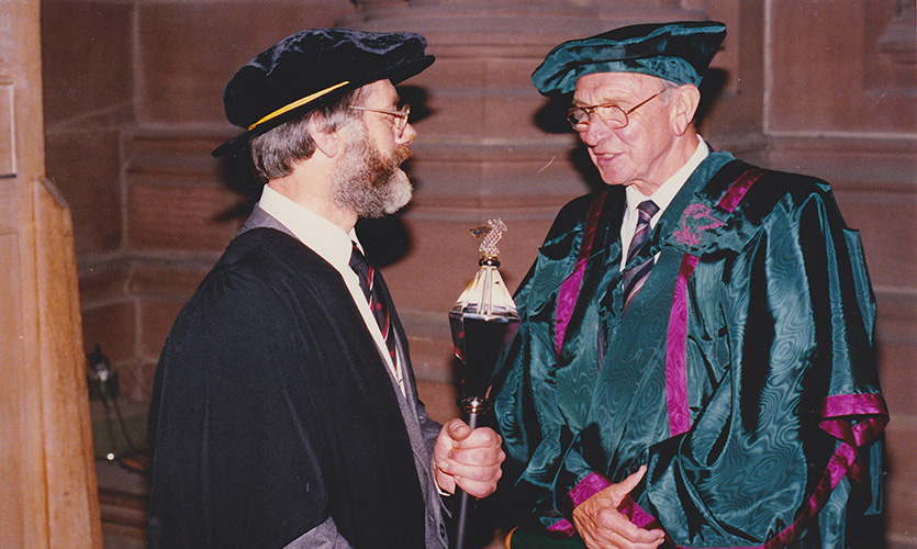 Arthur Hyatt stood on the right dressed in a cap and gown holding the mace in both hands as he talks to Honorary Fellow Jack Jones also dressed in a cap and gown 