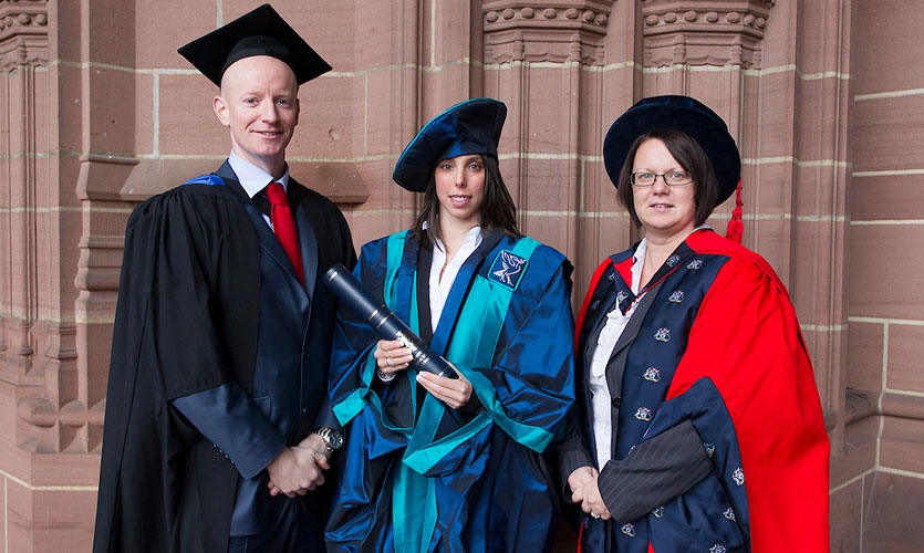 Beth wearing a cap and gown, holding her Honorary Fellowship outside of the Anglican Cathedral in Liverpool with LJMU staff Dave McDermott stood to her left and Professor Zoe Knowles stood on her right 