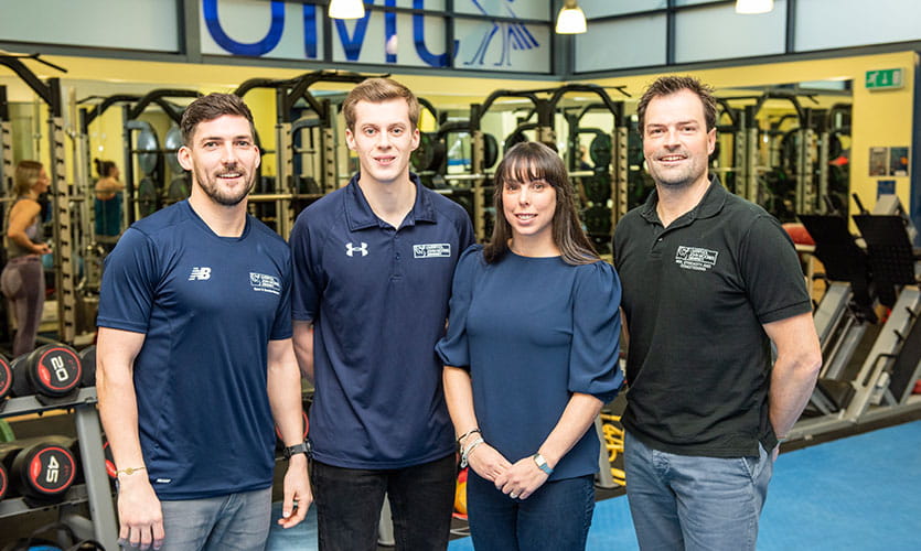 Beth stood in the university strength and conditioning gym with two members of LJMU staff stood to her left and one on her right hand side