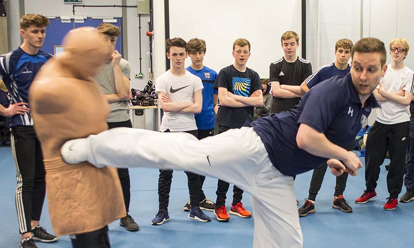 Carl is stood on a blue mat and his right leg is horizontal at 90 degrees kicking a plastic boxing dummy, stood in the background are a row of 8 male students .