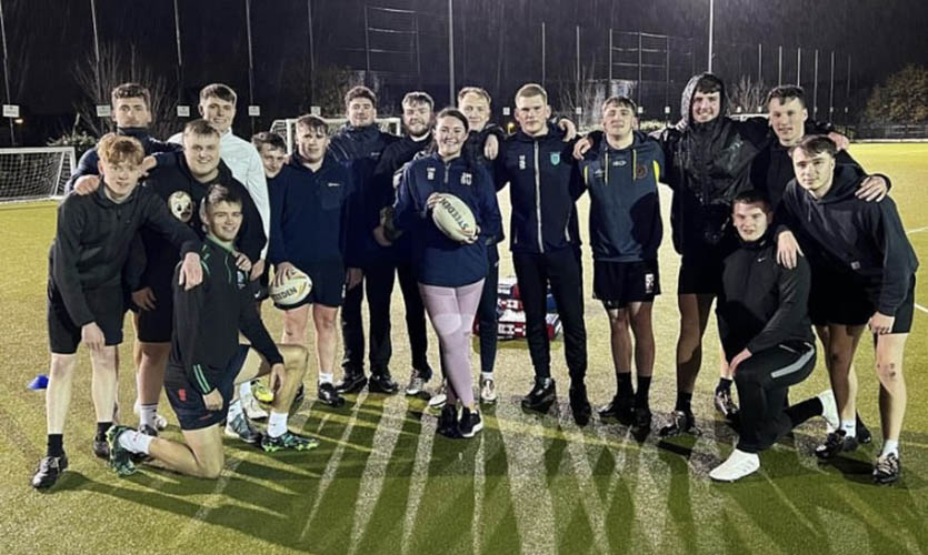 Clodagh is holding a rugby ball stood on a green astro turf pitch surrounded by male student rugby players dressed in navy sports shorts and t-shirts 
