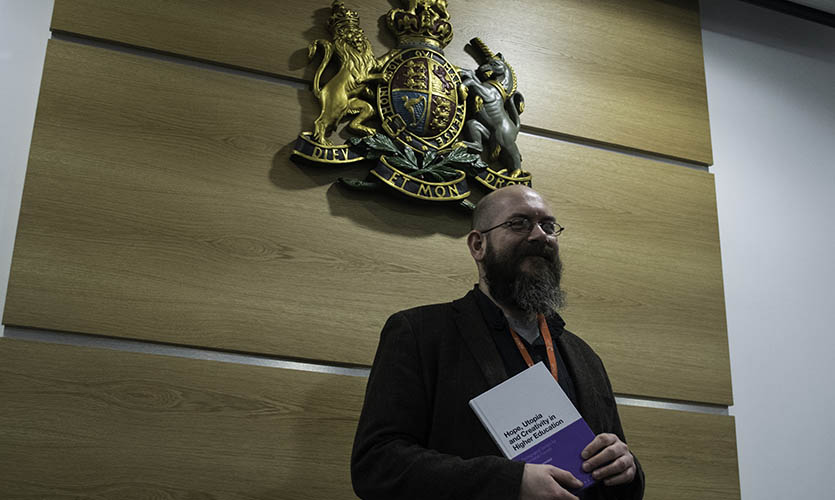 Craig is stood under the British Royal Coat of Arms in the Moot Room holding a book that he has written