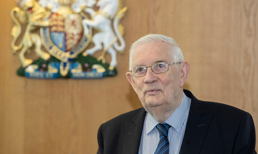 Head and shoulders photograph of David in the Moot Room, the Royal Coat of Arms of the United Kingdom is visible over his right shoulder on the wall in the background