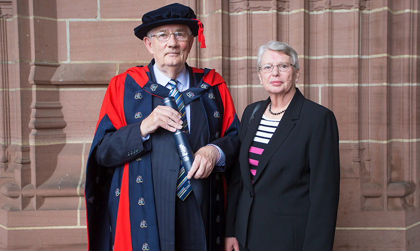 David dressed in a cap and gown after receiving his Honorary Fellowship stood outside of the Anglican Cathedral in Liverpool with his wife stood to his right hand side