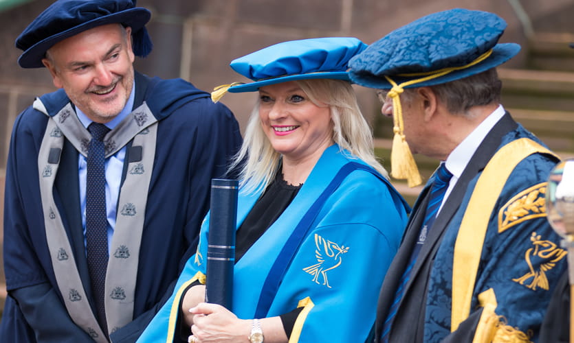 Denise is dressed in blue graduation robes and cap and can be seen from the waist up stood between two male LJMU members of staff also dressed in robes and caps.