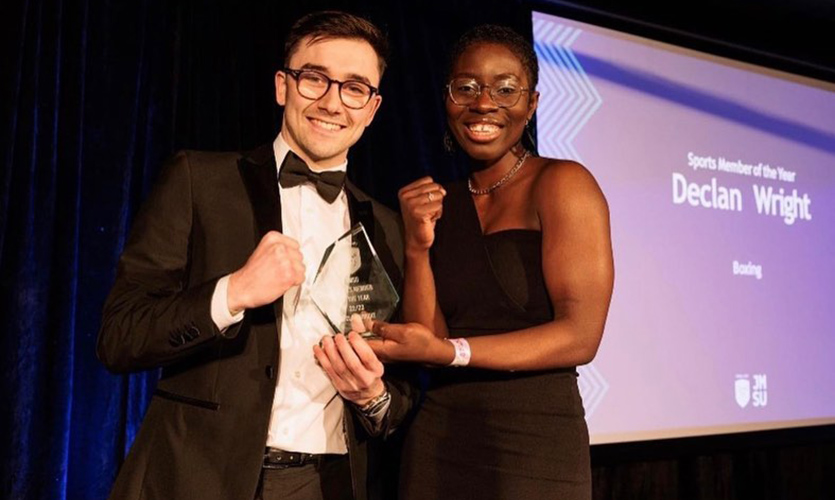 Dex is wearing black tie dress and is holding an award in his left hand and clenching his other hand into a fist, he is stood next to JMSU president Marie Hie who is handing the award over to him