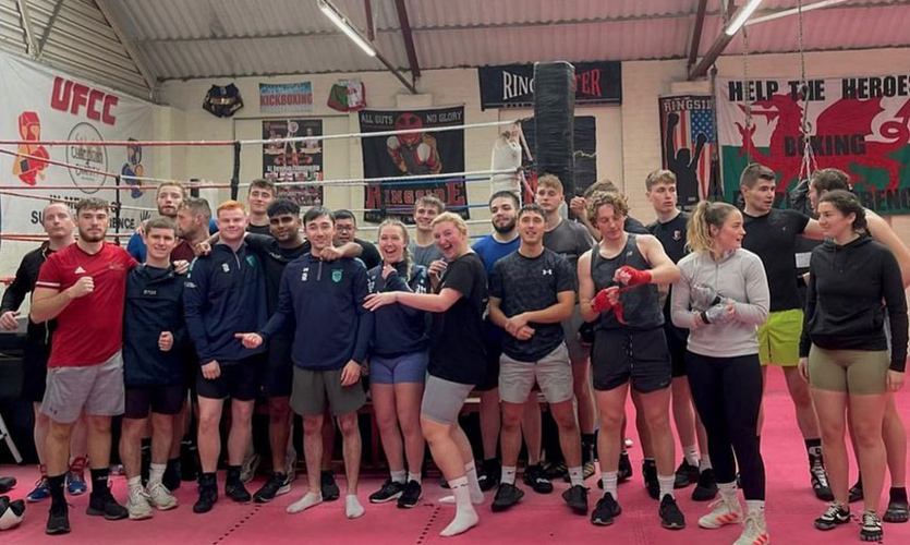 Dex is stood in the middle of a group of around twenty people, they are dressed in gym gear and stood in front of a boxing ring 