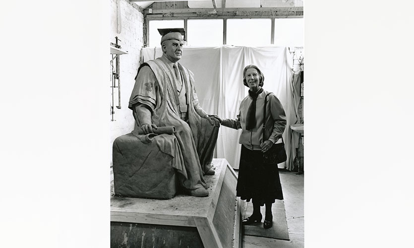A black and white photograph of the Henry Cotton statue being made in a workshop, Henry’s wife is stood to the right of it with her hand resting on the knee of the statue