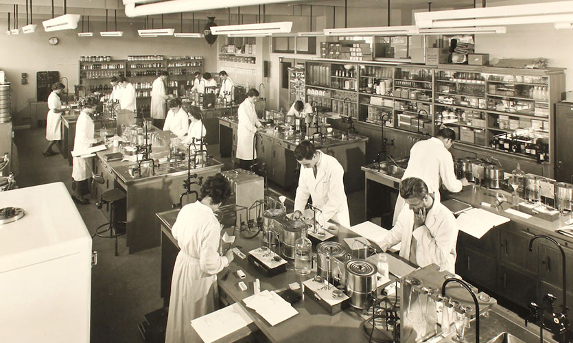 A black and white photograph showing a science laboratory in the 1960s with 16 people dressed in white lab coats undertaking experiments