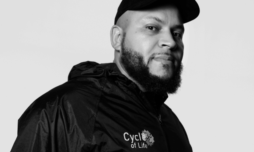 Black and white head and shoulders portrait photograph of Ibe looking to the camera wearing a black cap and dark coloured zip-up jacket with the words Cycle for Life embossed on the front left 