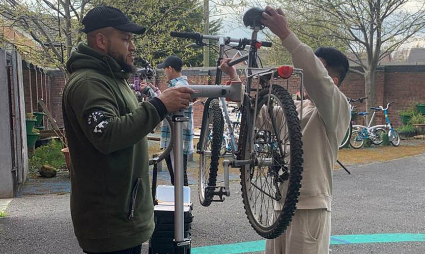 Ibe stood side-on holding a metal stand that has a bicycle clamped inside it and a teenage boy is stood the other side holding the bike seat and handlebars 