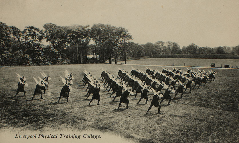 A black and white photograph showing 12 rows of women stood outside on a field all posed in the same position stretching one arm outwards and bending one knee