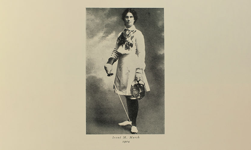 A black and white photograph of Irene Marsh stood wearing fencing clothing and holding a face guard in one hand and a fencing sabre in the other