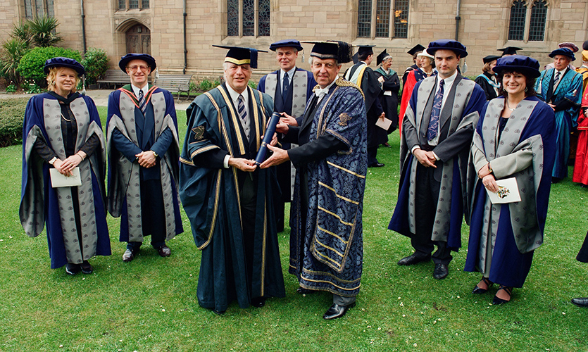 John is dressed in a ceremonial cap and gown and is surrounded by other men and women dressed in caps and gowns on the day of his installation as Chancellor