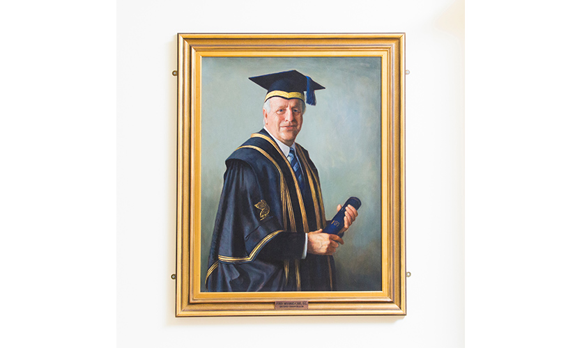A painted portrait of John Moores Junior he is painted wearing an LJMU cap and gown and holding a scroll in his hands