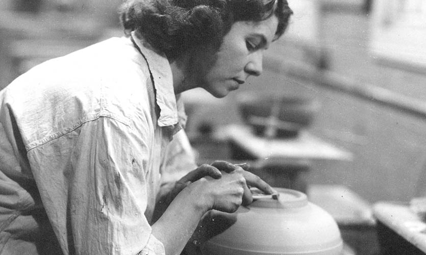 A black and white photograph of Julia taken side on, she is wearing a white loose-fitting shirt and is bent over working on a pottery wheel shaping the base of a bowl with her hands