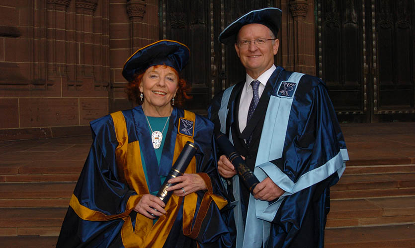 Julia is seen from the waist upwards wearing a graduation style satin blue cap and gown, she is holding a scroll in her hands
