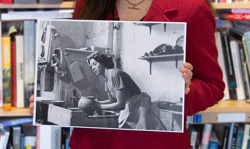 Dr Emma Roberts is stood in front of a bookcase and in front of her body she is holding a black and white photograph of Julia Carter Preston sculpting a pot on a kiln