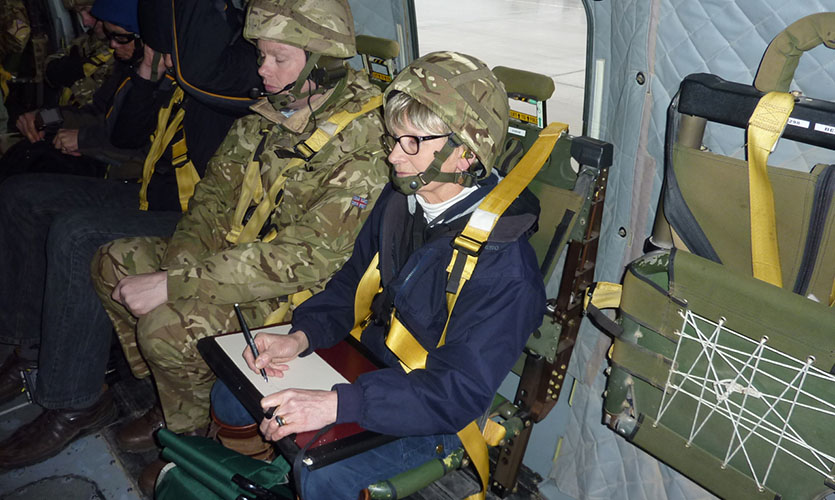 Julia is sat down in a military helicopter, wearing a camouflage helmet and holding a sketching pad and pen on her lap