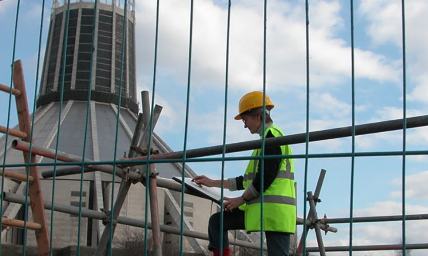 Julia is sat down on scaffolding holding her notepad and pen, in the construction site of the John Lennon Art and Design Building overlooking the Metropolitan Cathedral in Liverpool 