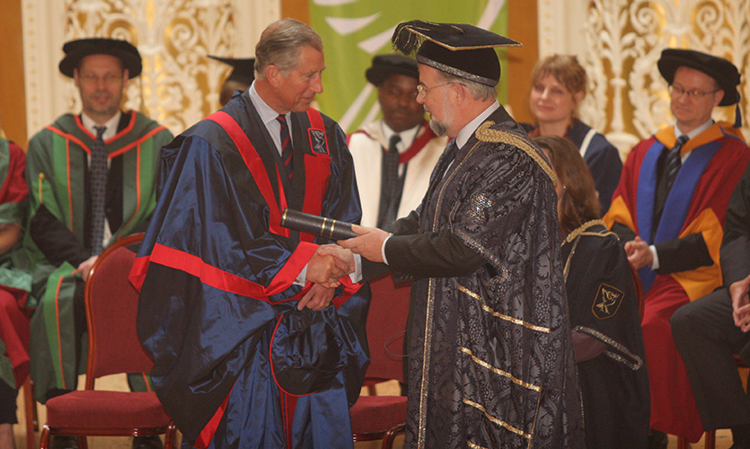 The Prince of Wales is dressed in blue graduation style robes and is being handed a scroll on a stage by another man wearing similar graduation style robes and a hat
