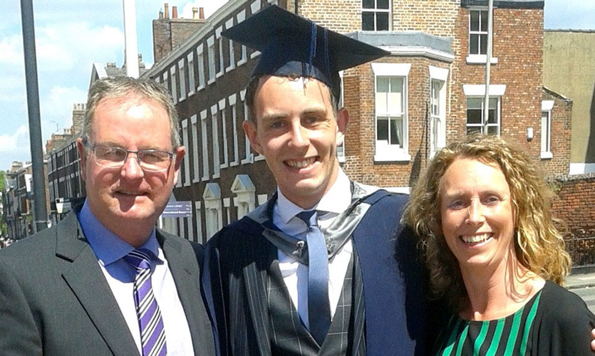 Photograph of Nick stood in between his parents on his graduation day, Nick is smiling and wearing a graduation cap and gown over a three-piece suit 