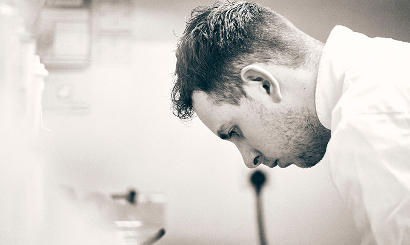 A sepia filtered photograph taken close and side on of Nick in a kitchen, he has a short back and sides haircut and stubble on his face and is looking downwards