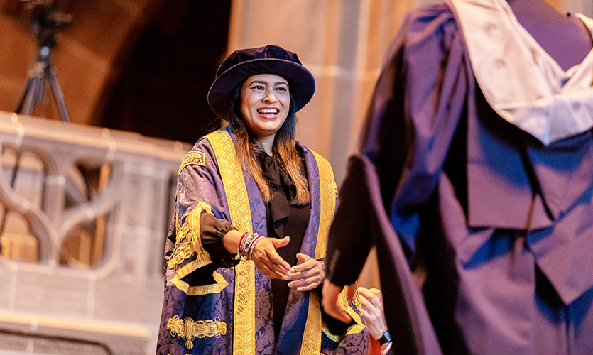 Nisha is smiling and is dressed in a ceremonial cap and gown with her hand outstretched ready to shake the hand of an LJMU graduate crossing the stage in Liverpool Cathedral