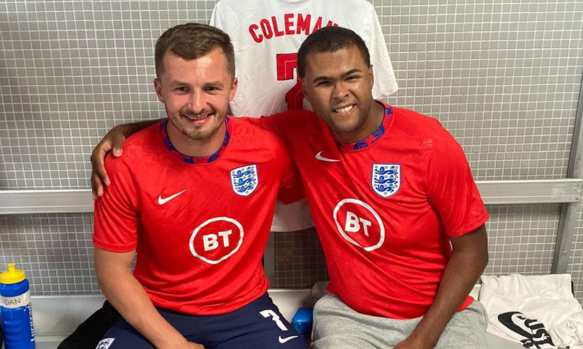 Rainbow is sat on a bench in a football changing room wearing a red England branded football shirt with his arm extended around a fellow male blind football player who is sat to his left