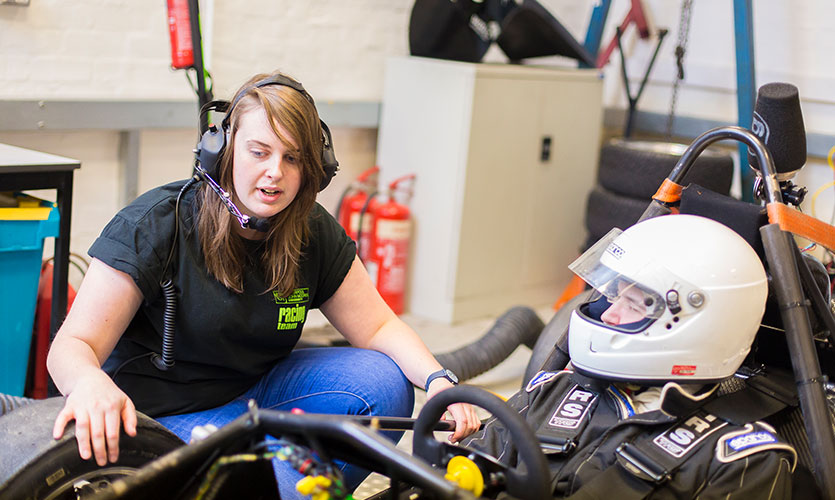 Rhian is wearing a headset with a microphone crouched down over a racing car which has a person sat at the steering wheel wearing a helmet