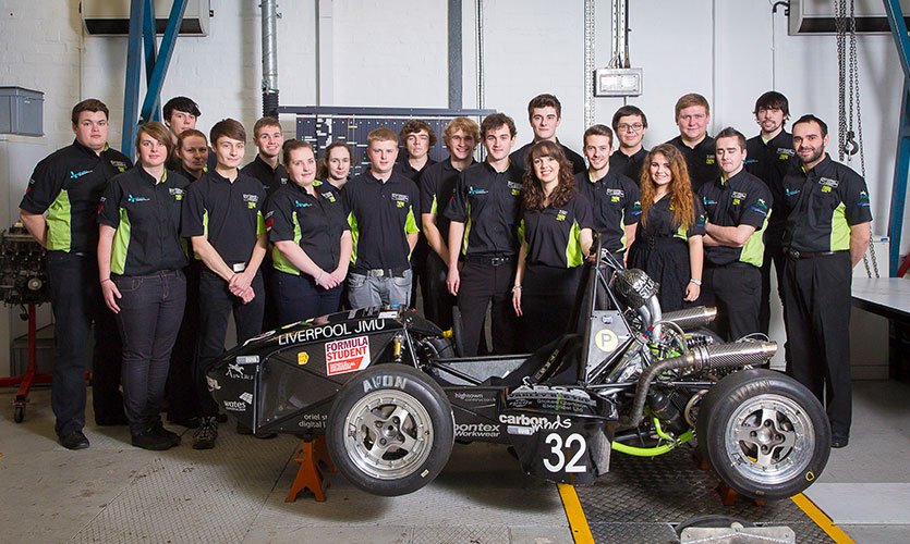 21 students all dressed in the same black and green shirt are stood behind a racing car parked sideways with two wheels visible at the front
