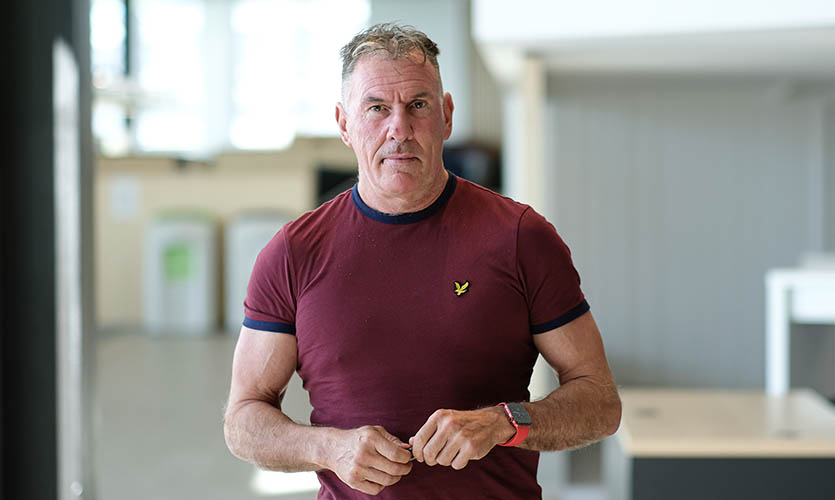 Robert pictured from the waist up, standing in a university building wearing a maroon t-shirt with his arms clasped in front of him
