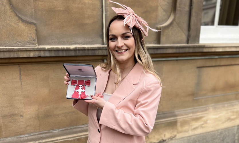 Sam is dressed in a pale pink trouser suit and is stood outside of a brick building holding her MBE medal in an opened bow with both of her hands