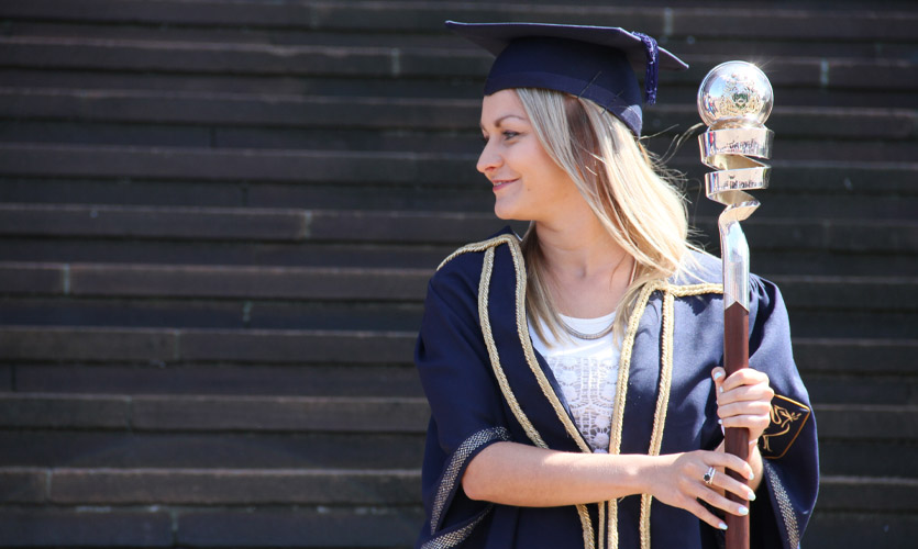 Sam is wearing a graduation cap and gown and is looking to the side; she is picture from the waist up and is holding a silver and wooden mace in her hands