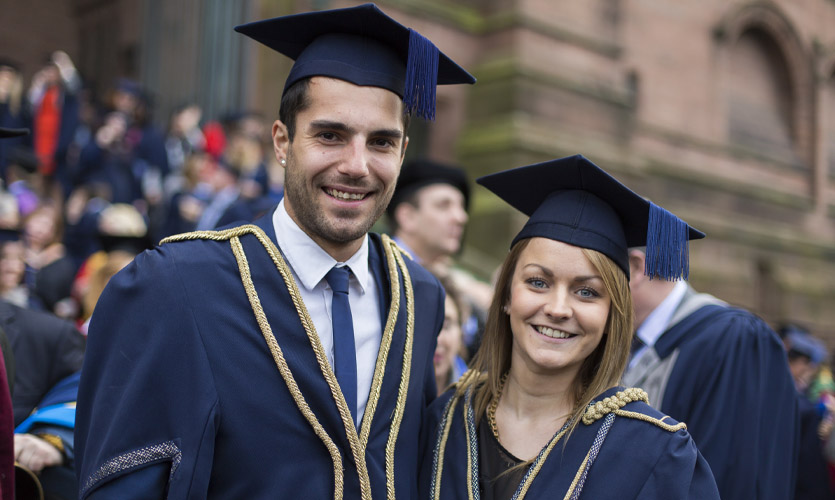 Head and shoulders of Sam who is wearing a navy-blue graduation cap and gown, she is stood next to a man, and they are both smiling