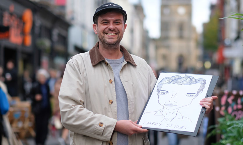Sean can be seen from the waist up wearing a grey t-shirt, beige jacket and black cap, stood on Bold Street in Liverpool city centre holding a large white canvas in his hands featuring a black line drawing of popstar Harry Styles’ face