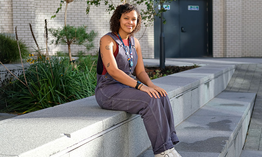 Shaquita is sat down on a concrete bench outside the Student Life Building, she is smiling and has her hands placed on her knees, she is wearing denim dungarees and white chunky soled boots 