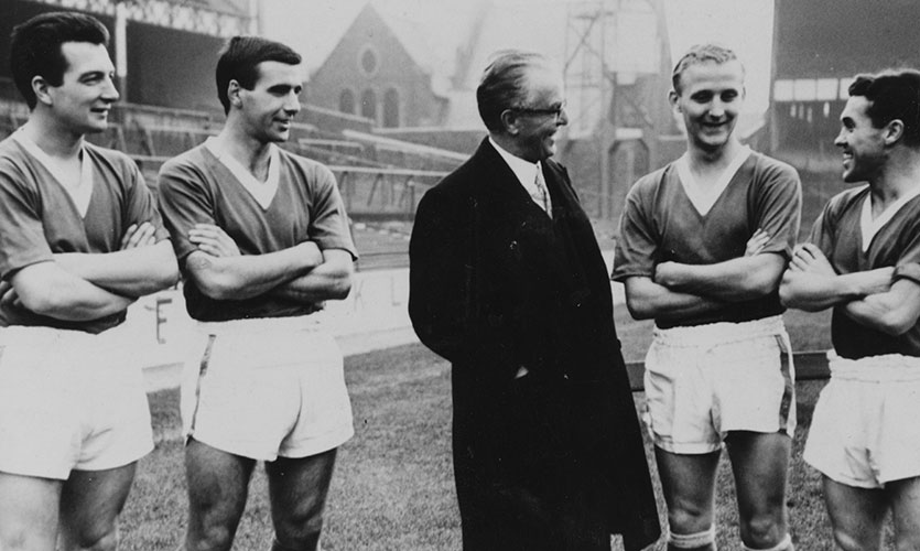 A black and white photograph of Joh stood on a football pitch with two players stood either side of him