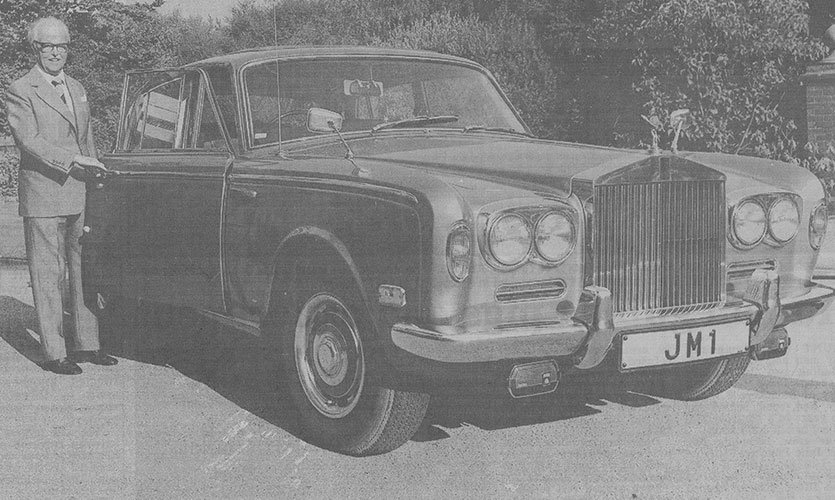A black and white photograph of John stood to the left of a Rolls Royce car featuring a registration plate with the letters J M 1