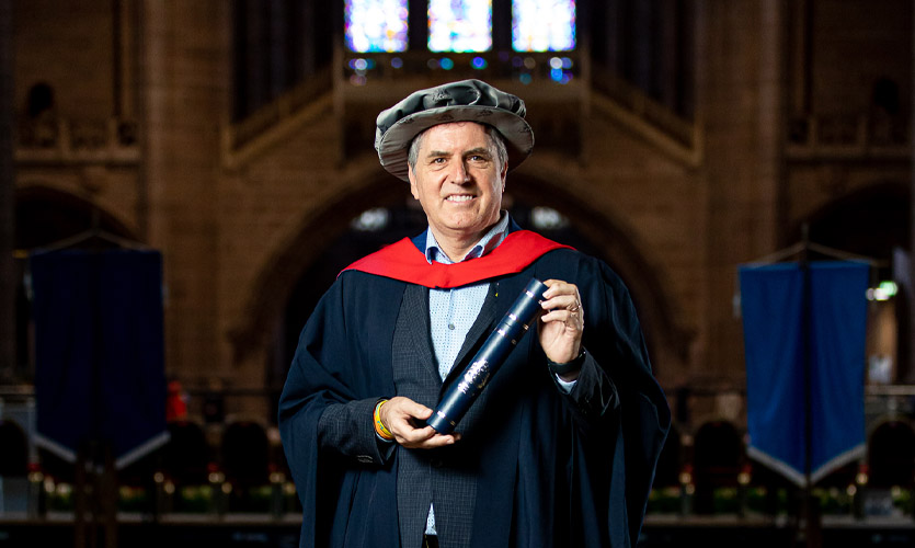 Steve dressed in a cap and gown holding a scroll in his hands with the Liverpool Cathedral large stained glass window seen in the background