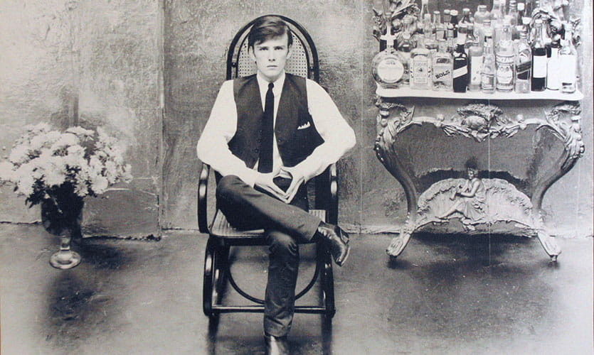 A black and white photograph of Stuart sat in a wooden chair with his legs crossed there is a vase of flowers on the floor on the left of him and a table with a mirror and glass bottles to the right