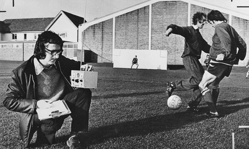 A black and white photograph of Tom couched down on a football pitch holding science equipment with two people playing football in the background